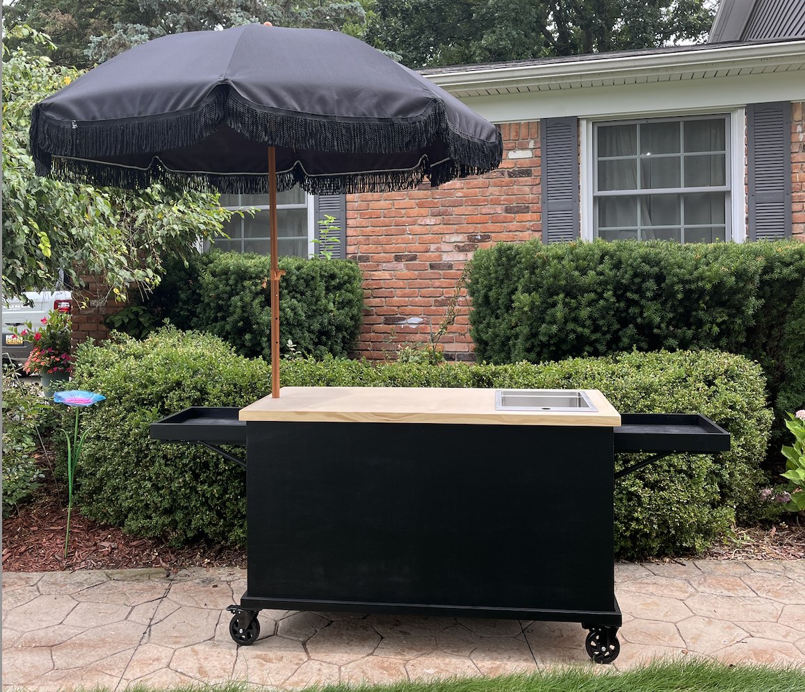 Black coffee cart with a natural wood top, a stainless drop-in sink, fold-out side trays and a black fringed parasol
