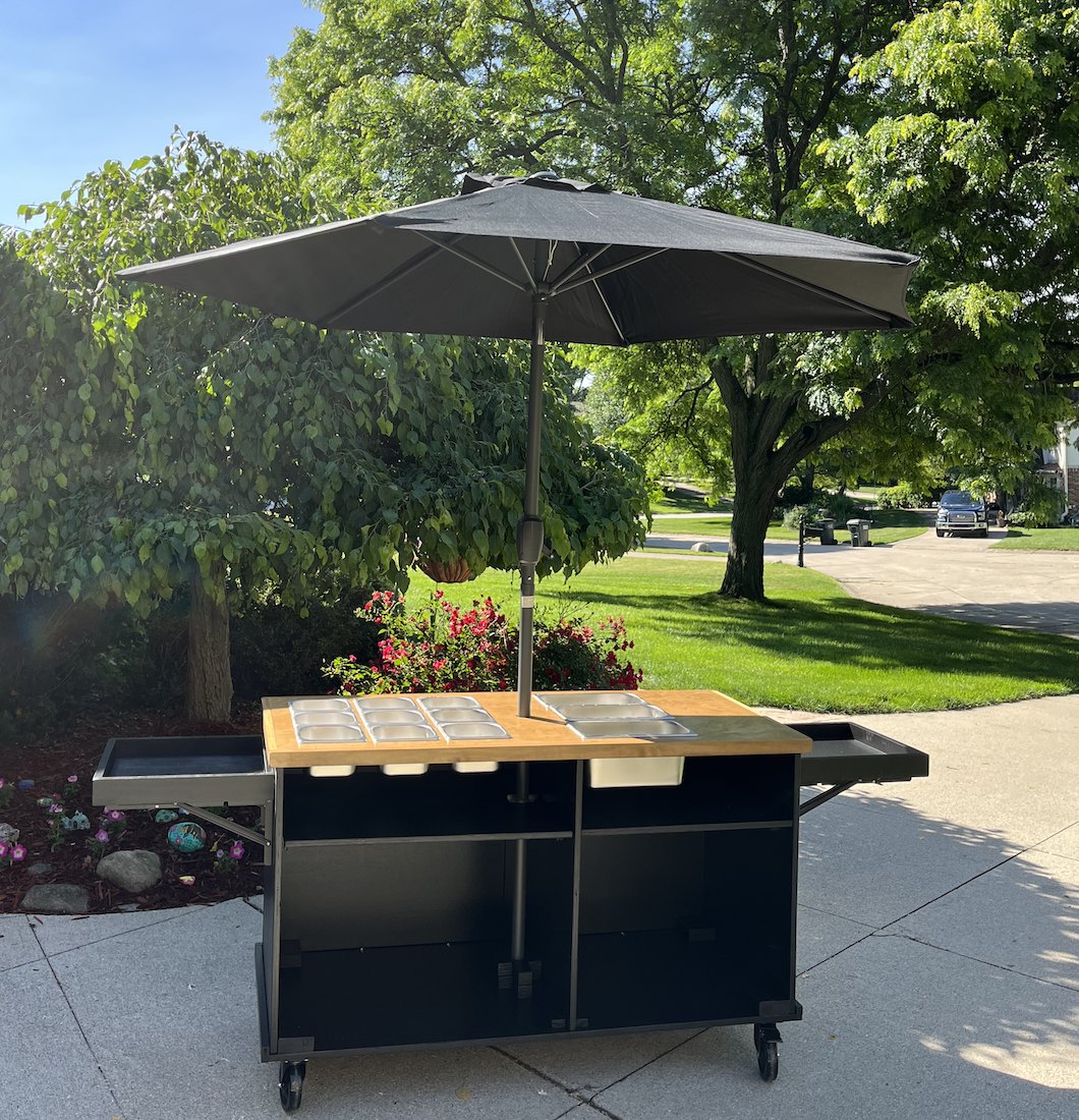 The same black cart from an angle, open to show the wood top with food wells, fold-out side shelves on both sides, and the center umbrella