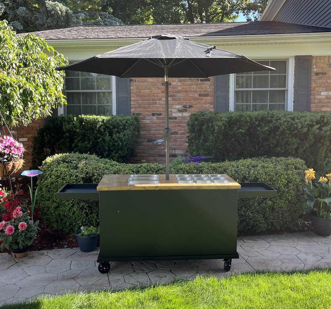 New black grazing cart with a signature wood top and food wells, with the umbrella set through the center, shown from the front outdoors