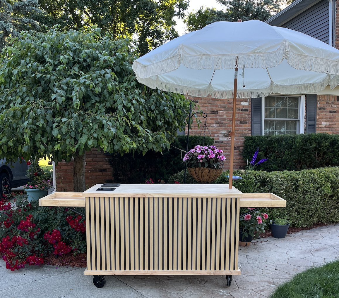 New slatted-wood coffee cart: a natural wood panel wrapping all the way around with a signature wood top, fold-out wood side shelves and a cream fringed umbrella, set up outdoors