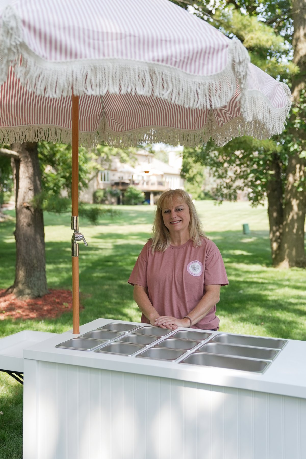 Laura, founder of Woodshop Joy, smiling behind a white handcrafted wells cart with a pink-striped fringed umbrella